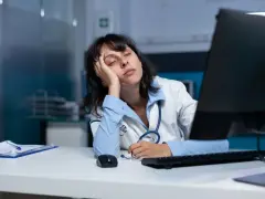 Zmęczona lekarka opiera głowę na dłoni, zasypiając przy biurku. Długie godziny pracy w nocy dają się we znaki.