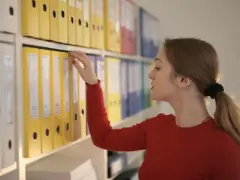 Kobieta szuka dokumentów, być może dotyczących ewidencji czasu pracy umowa zlecenie, wśród kolorowych segregatorów na półce.